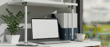 Pencerenin yanındaki modern offie çalışma alanı dizüstü bilgisayarlı boş ekran maketi, dekor bitkisi, kahve fincanı ve ofis malzemeleri. 3d görüntüleme, 3d illüstrasyon