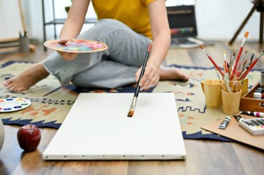 Genç Asyalı kadın stüdyo zemininde oturuyor, resim yapıyor, boya fırçası ve akrilik renkle boş tuvali boyuyor..