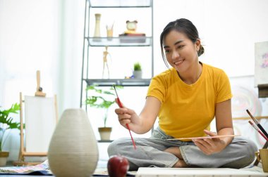 Yetenekli ve yaratıcı genç Asyalı kadın tasarımını boya fırçasıyla ölçüyor, ev stüdyosunda bazı sanat işleri yapıyor..