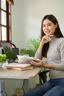 Portre, çekici ve çekici Asyalı genç bayan yönetici ya da günlük kıyafetli iş kadını ofis masasında oturuyor, elleri çenesinde, kameraya bakıp gülümsüyor..