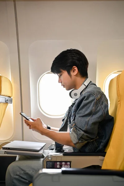 Hipster young Asian male traveler or passenger in an economy seat, using his smartphone to chat with someone during the flight.