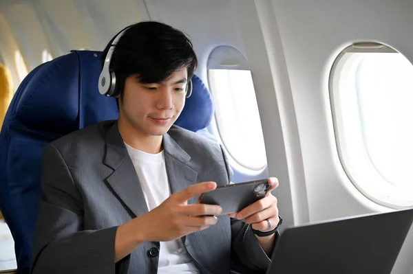 stock image Handsome young Asian businessman or male passenger wearing wireless headphone and playing a mobile game during the flight.