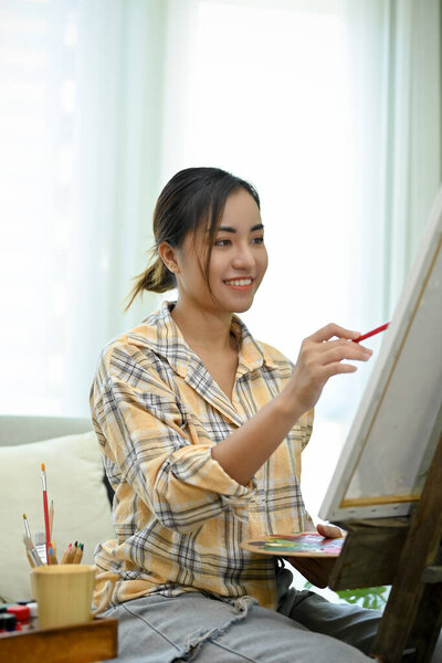 Portrait, Cheerful young Asian woman artist sitting in front of easel in her workshop, making picture, painting with acrylic color. creativity hobby concept.