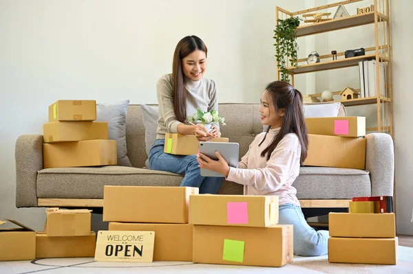 stock image Two young Asian female small e-commerce business owners working, checking or orders on the website and preparing shipping boxes together in living room.