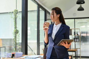 Güzel ve muhteşem Asyalı iş kadını ofisinde hayal kurarken kahvesini yudumluyor..