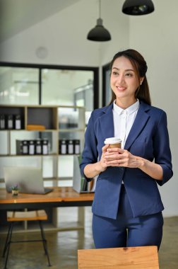 Portre, çekici ve kendinden emin bin yıllık Asyalı iş kadını ya da takım elbiseli kadın patron elinde kahve fincanıyla ofisinde duruyor..