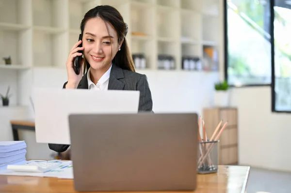 stock image Charming young Asian businesswoman or female secretary talking on the phone with business partner at her desk.