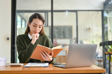 Kararlı ve profesyonel Asyalı iş kadını veya kadın finans çalışanı kitap okuyor, bir ders kitabında bilgi araştırıyor, masasında çalışıyor..