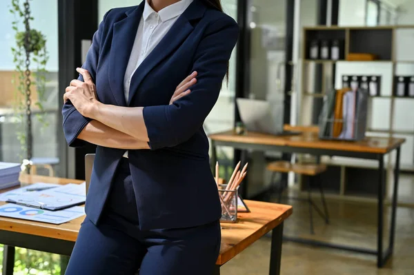 Professional and successful millennial businesswoman in formal business suit arms crossed and stands in her modern office. cropped image