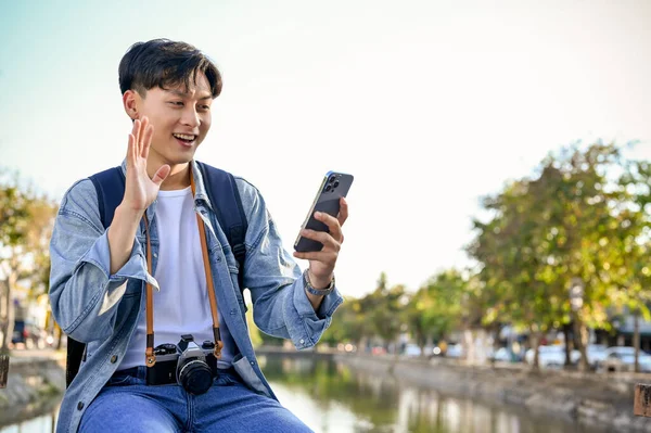 Yakışıklı ve mutlu Asyalı genç erkek gezgin gülümsüyor, el sallıyor, akıllı telefondan arkadaşıyla video görüşmesi yapıyor ve şehirde gezinmeyi seviyor..