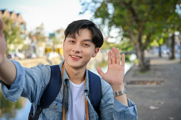 Handsome and attractive young Asian male traveler or travel blogger smiling, waving hand for greeting, looking at camera.