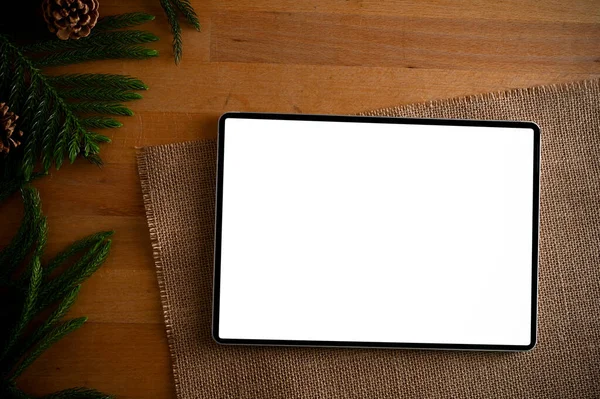 Top view, A digital tablet white screen mockup for display your graphic ads is on a wood tabletop with green tree branch.