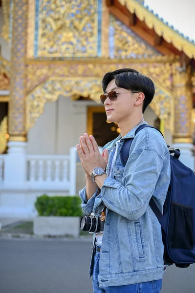 Portrait, Handsome and attractive young Asian male traveler putting his hands together in a prayer position in front of the beautiful Thai\'s temple. tourist, Asia traveling concept