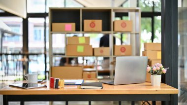 Dizüstü bilgisayar, tablet, ofis malzemeleri, karton kutular ve paketleme ekipmanlarıyla dolu bir paketleme odasında ofis masası..