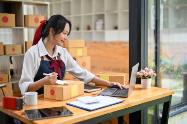 Güzel ve çekici Asyalı genç bayan küçük online işletme sahibi posta kutularını hazırlarken bilgisayarından siparişlerini kontrol ediyor..