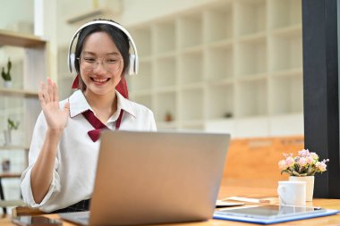 Güzel ve mutlu Asyalı genç bir kadın çevrimiçi toplantıya ya da webinar 'a katılır, elini sallayarak takımını selamlar ya da dizüstü bilgisayarla selam verir..