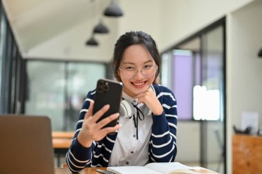 Güzel, genç Asyalı bir bayan masasında oturuyor telefonuna bakıyor ve gülümsüyor, akıllı telefonunu kullanıyor, blog okuyor, sosyal medyada kayıyor, selfie çekiyor..