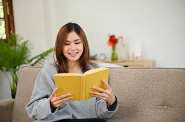 Güzel ve çekici genç Asyalı kadın rahat oturma odasında dinlenirken kitap ya da roman okumaktan hoşlanıyor..