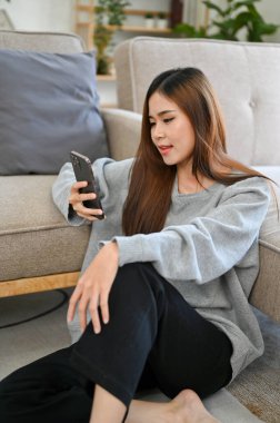 Rahat ve rahat genç Asyalı bir kadın, günlük kıyafetleriyle, hafta sonunu evde geçirir, rahat oturma odasında oturur ve telefonunu kaydırır..