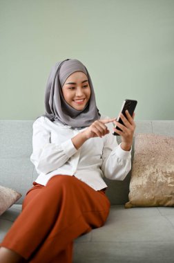Portre, güzel Asyalı genç Müslüman kadın tesettürlü akıllı telefonunu arkadaşlarıyla sohbet etmek için kullanıyor ya da oturma odasında dinlenirken internetten alışveriş yapıyor..