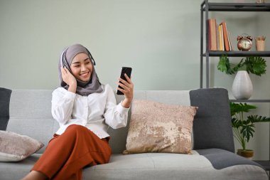 Bin yıllık çekici Asyalı Müslüman kadın tesettür ve kulaklık takıyor cep telefonunu kullanıyor, oturma odasında dinlenirken en sevdiği şarkıyı dinliyor..