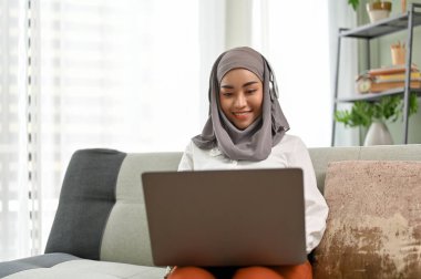 Profesyonel ve çekici Asyalı Müslüman iş kadını tesettür ve kulaklık takıyor, evden çalışıyor, dizüstü bilgisayar kullanıyor, online bir toplantıya katılıyor veya webinar.