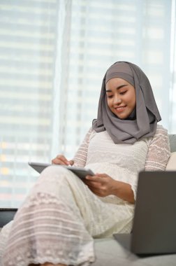 Portre, güzel Asyalı Müslüman kadın oturma odasında dinlenirken dijital tablet kullanıyor..
