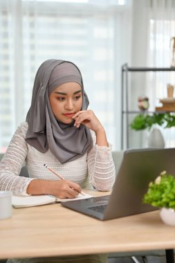 Portre, düşünceli Asyalı genç Müslüman kız üniversite öğrencisi okul ödevini düşünerek evde dizüstü bilgisayarıyla ödev yapıyor..