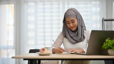 Çekici ve profesyonel Asyalı Müslüman iş kadını ofiste çalışan, yazı yazan ya da not alan tesettürlü.