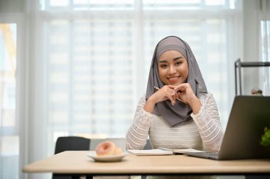 Muhteşem ve güzel Asyalı Müslüman iş kadını ya da tesettürlü kadın yönetici ofis masasında oturuyor, gülümsüyor ve kameraya bakıyor..