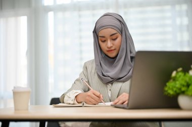 Profesyonel ve güzel Asyalı Müslüman iş kadını ya da iş kadını projesine konsantre olup planını masasındaki bir kitaba yazıyor..