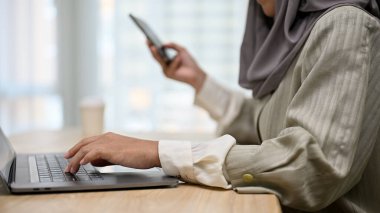 Kesilmiş görüntü, profesyonel Asyalı Müslüman iş kadını ya da ofiste çalışan kadın yönetici, dizüstü bilgisayar kullanıyor, klavyede telefon tutarken daktilo kullanıyor..