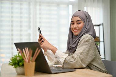 Çekici ve muhteşem Asyalı Müslüman iş kadını veya tesettürlü kadın yönetici ofis masasında oturuyor, akıllı telefon kullanıyor, gülümsüyor ve kameraya bakıyor..