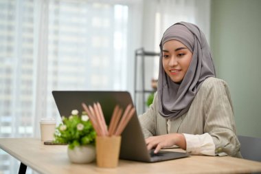 Çekici ve profesyonel Asyalı Müslüman iş kadını ofiste çalışırken tesettürlü, dizüstü bilgisayar kullanıyor..
