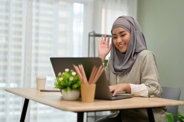 Çekici Asyalı Müslüman iş kadını ya da tesettürlü kadın ofis çalışanı çevrimiçi toplantı yapıyor ya da ekibiyle el sallayarak iş arkadaşını selamlıyor..