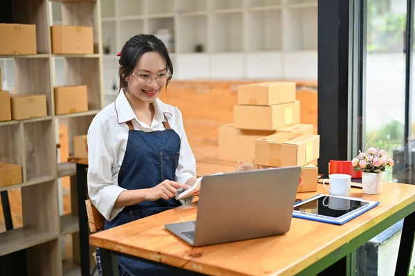 Successful young Asian female small online business owner working in her packing room, replying customer\'s question on the website via laptop, using calculator to check the price.