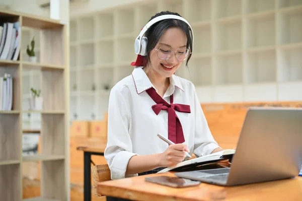 stock image Charming young Asian female student wearing eyeglasses and headphones, doing homework, writing something in her school notebook while taking an online class.