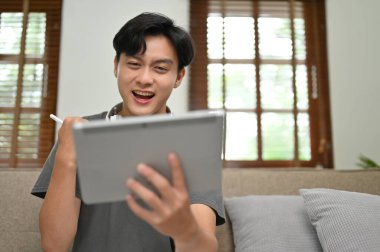 Heyecanlı ve neşeli Asyalı genç adam tablet ekranına bakıyor, sevinçten bağırıyor, beklenmedik bir haber aldı..
