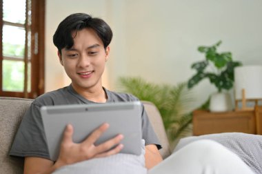 Yakışıklı ve mutlu Asyalı genç adam kanepeye oturur ve dijital tablet dokunmatik ped kullanır. Teknoloji konsepti