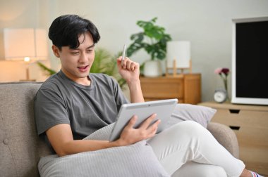 Yakışıklı Asyalı genç adam hafta sonunun tadını çıkarıyor. Oturma odasında dinlenirken taşınabilir dijital tablet kullanıyor..