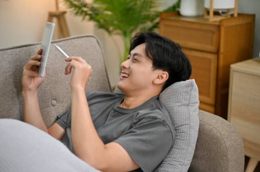 Neşeli ve yakışıklı Asyalı genç adam oturma odasında rahat koltuğunda uzanıyor, dijital tablet kullanıyor, dokunmatik pedinde oyun oynuyor..