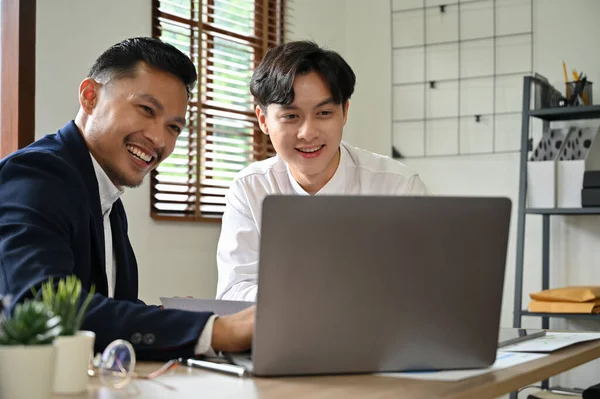 Cheerful and smart Asian businessman working with his coworker in the office. Male marketing employee shows his project plan on the laptop screen to his colleague.