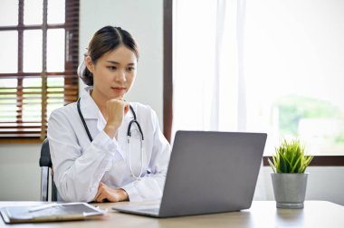 Profesyonel ve kendine güvenen genç Asyalı doktor ya da beyaz önlüklü ve steteskoplu doktor çalışma masasında oturuyor, dizüstü bilgisayarına bakıyor, özel ameliyat vakalarını düşünüyor ve planlıyor..