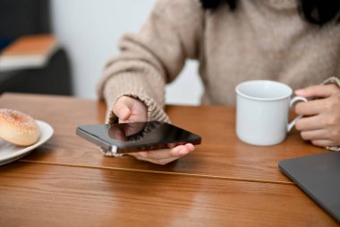 Rahat süveter giyen Asyalı bir kadın sabah kahvesini yudumlarken akıllı telefondan e-postalarını kontrol ediyor. kırpılmış atış