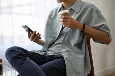 Yakışıklı, genç Asyalı bir adam kafeteryada dinlenirken akıllı telefonunu kullanıyor ve kahvesini yudumluyor. kırpılmış resim