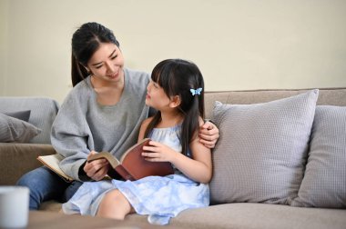 Güzel ve sevimli Asyalı anne kitap okuyor ya da küçük tatlı kızına komik bir masal anlatıyor oturma odasında dinlenirken. Mutlu aile kavramı