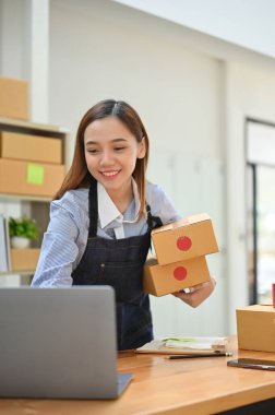 Portre, güzel ve başarılı Asyalı kadın e-ticaret girişimi paketleme odasında çalışıyor, dizüstü bilgisayardaki siparişleri kontrol ediyor ve nakliye paketlerini hazırlıyor..