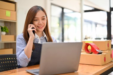 Milenyum Asyalı güzel bayan mağaza sahibi veya işçisi telefonda tedarikçisiyle hammadde siparişi verirken dizüstü bilgisayar ekranına bakıyor. küçük çevrimiçi iş konsepti