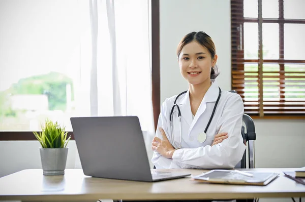 迷人而自信的年轻亚洲女医生坐在她的写字台前 双臂交叉 微笑着对着相机 专科医生 从业人员 — 图库照片