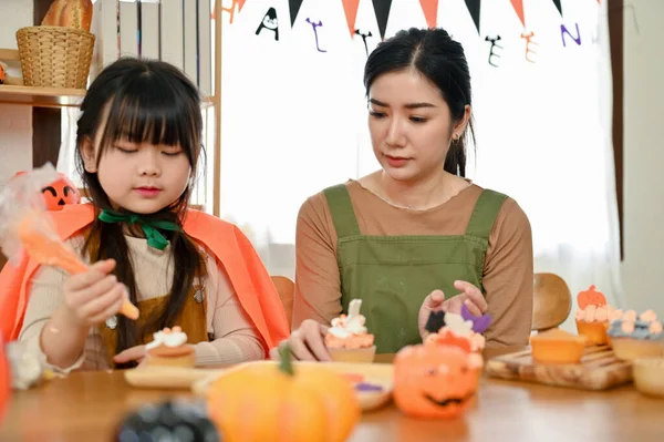 Lovely and kind Asian mom and her little cute daughter making a Halloween cupcake in the kitchen together. Happy family and Halloween day